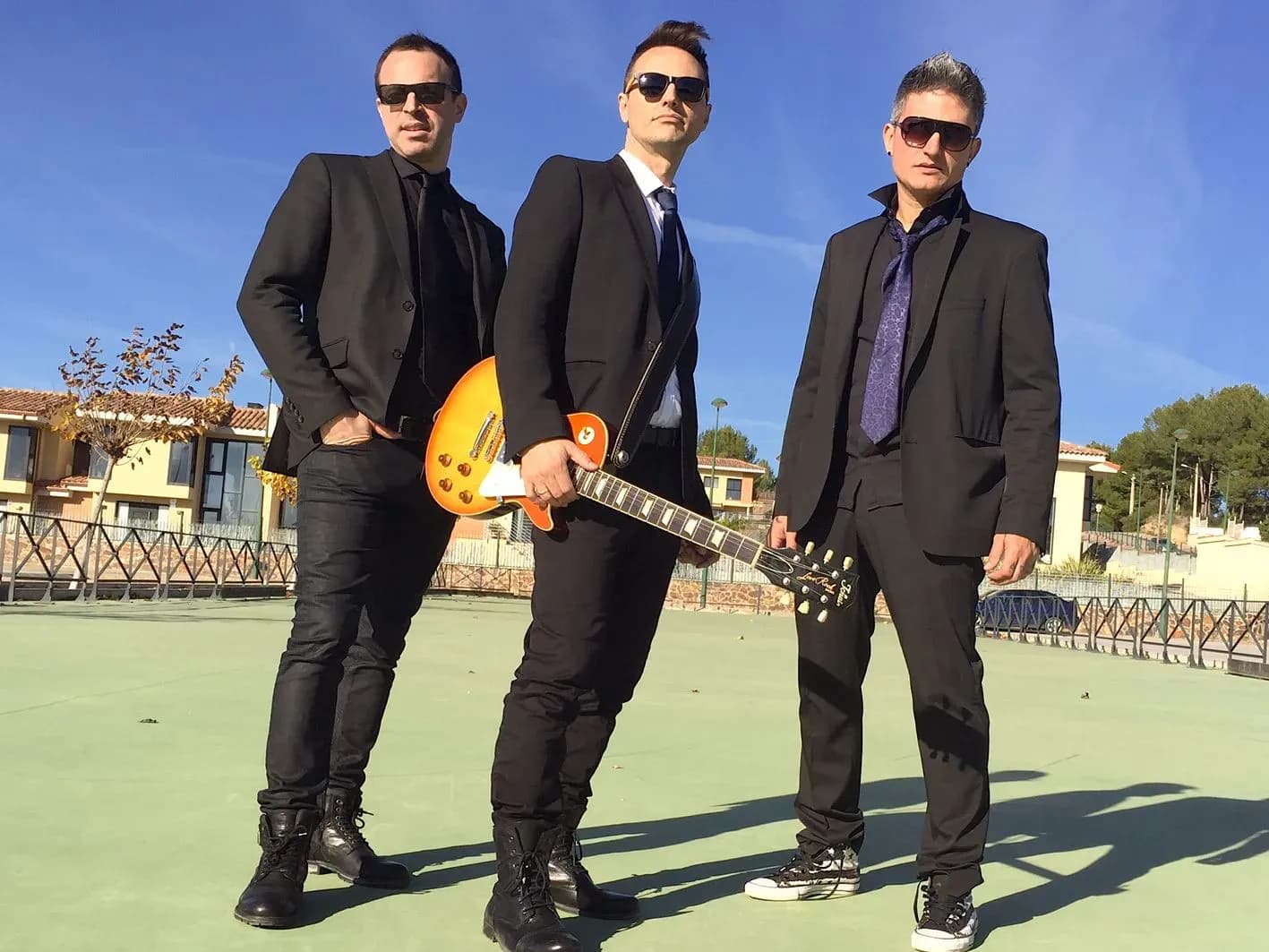 Mind Trick en formato trío con trajes negros y guitarra Les Paul naranja posando en pista deportiva bajo cielo azul