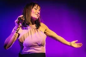 Cantante de Miris Way con microfono sobre escenario iluminado con luz violeta y brazos extendidos durante actuacion