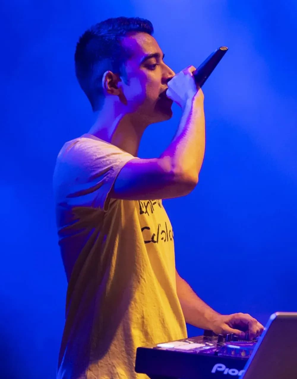 Quim Martínez con camiseta amarilla cantando al micrófono junto a la mesa Pioneer bajo luz azul de escenario