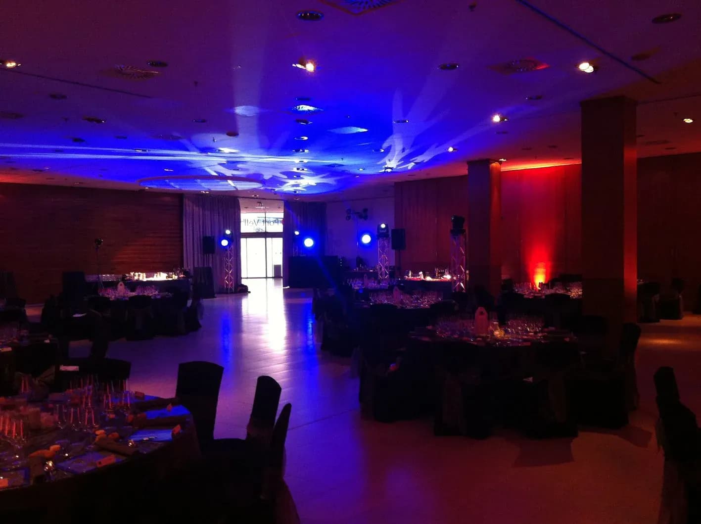 Salón de banquete con iluminación arquitectónica azul cenital y ámbar lateral, mesas vestidas y altavoces para evento corporativo