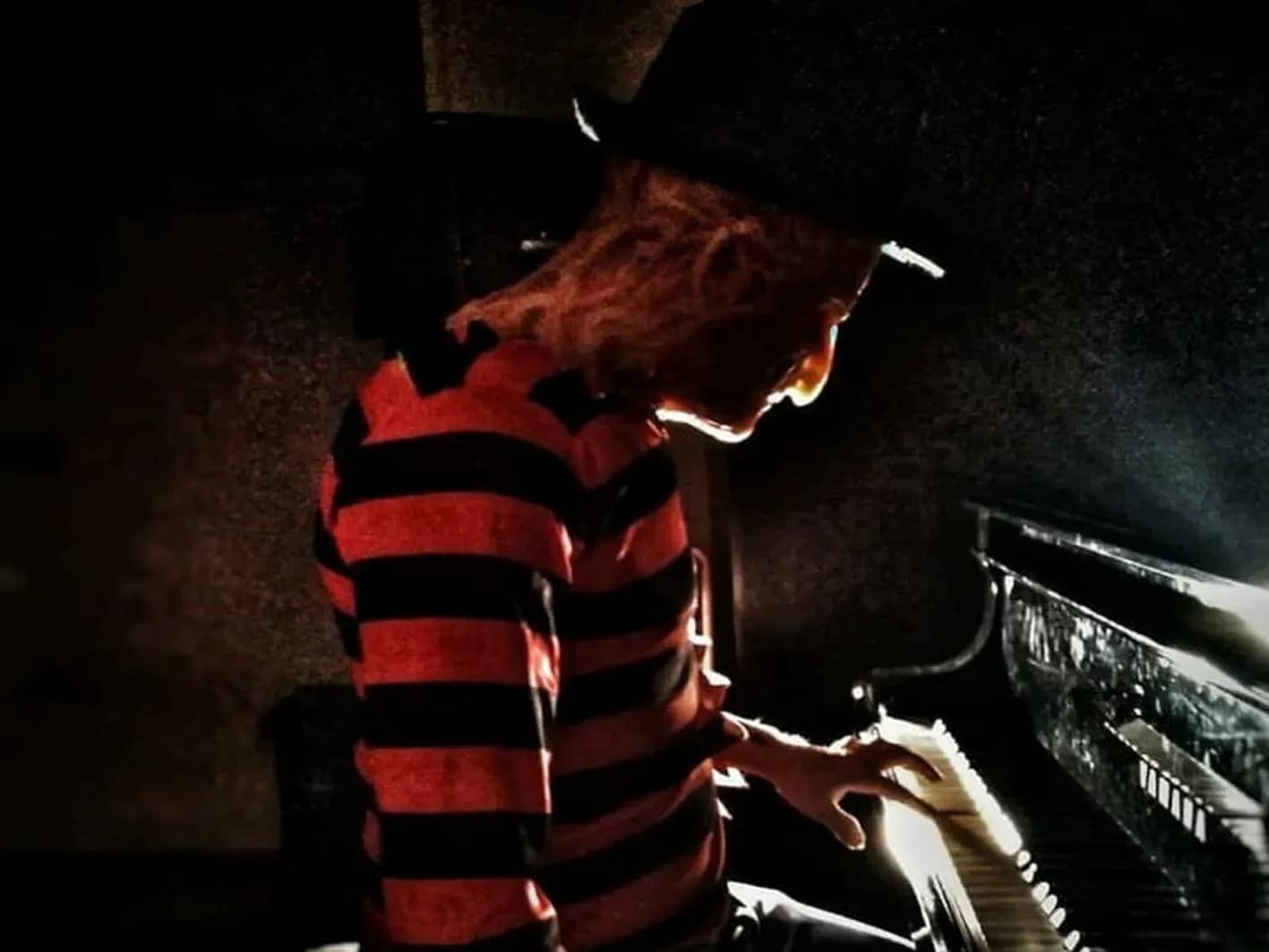 Actor caracterizado como Freddy Krueger con jersey de rayas rojinegras y sombrero tocando el piano en penumbra, escena de terror
