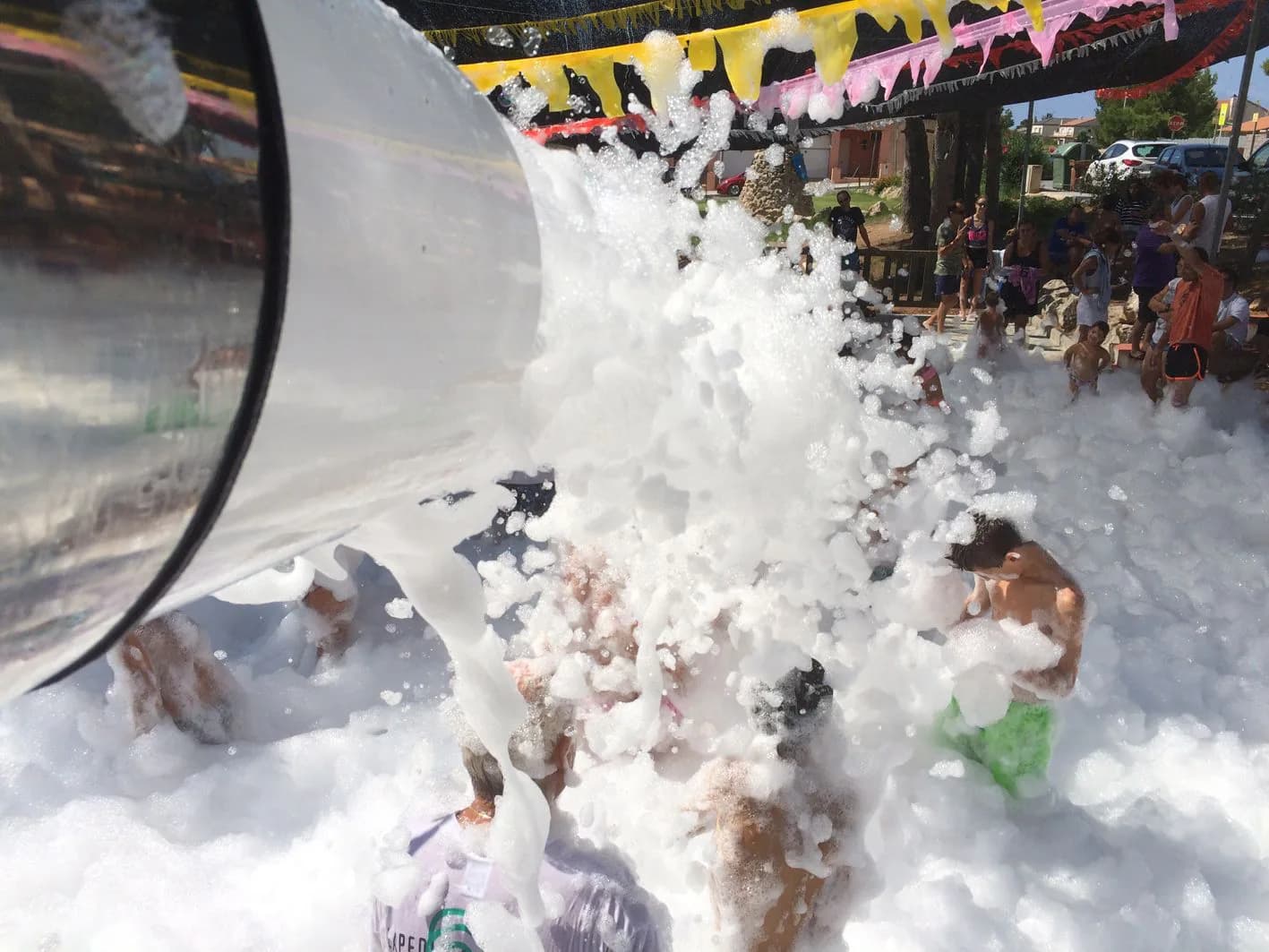 Cañón de espuma lanzando chorro blanco sobre niños bañándose entre montañas de espuma bajo guirnaldas de plástico