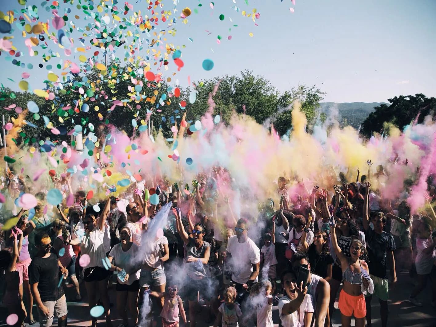 Multitud lanzando polvos Holi de colores y confeti al aire al atardecer con árboles y montañas al fondo, fiesta india