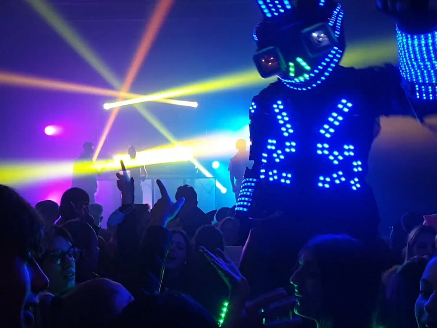 Robot LED gigante azul con casco iluminado caminando entre el público bajo láseres amarillos en discoteca