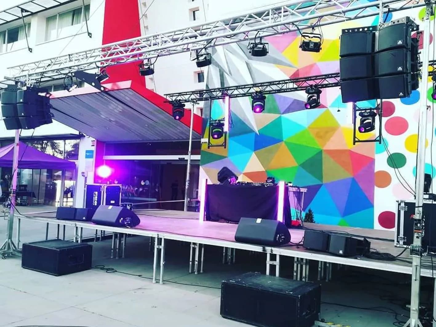 Escenario al aire libre con line array Adamson, focos LED, truss y cabina de DJ ante mural geométrico de colores