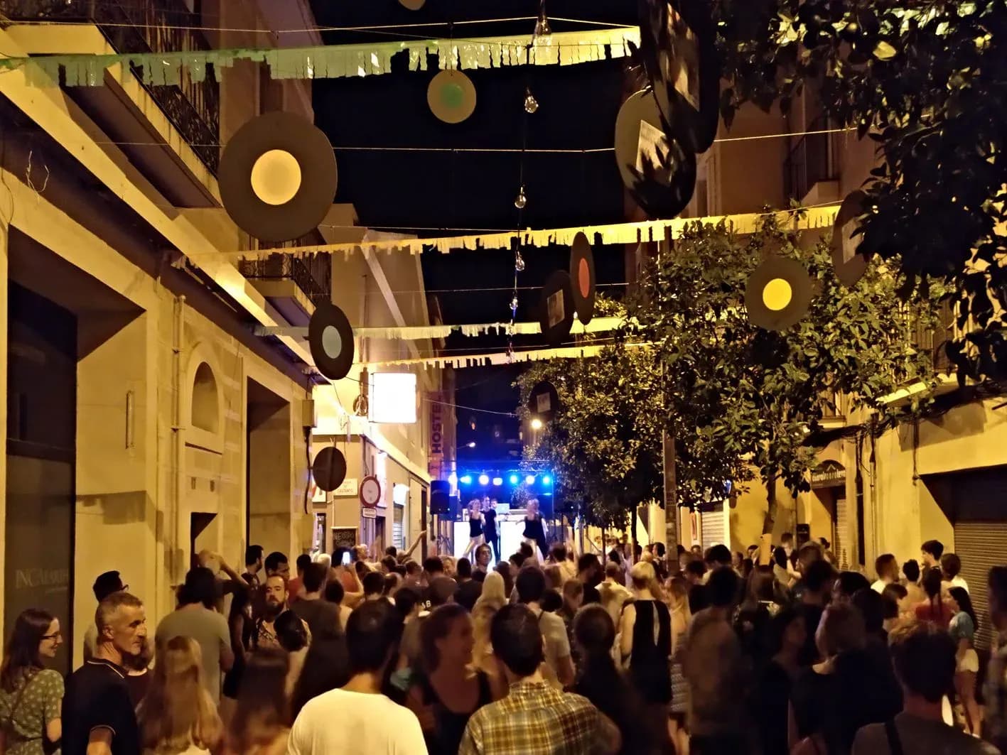 Calle de fiesta mayor con guirnaldas circulares de colores colgando entre fachadas y publico bailando ante escenario nocturno