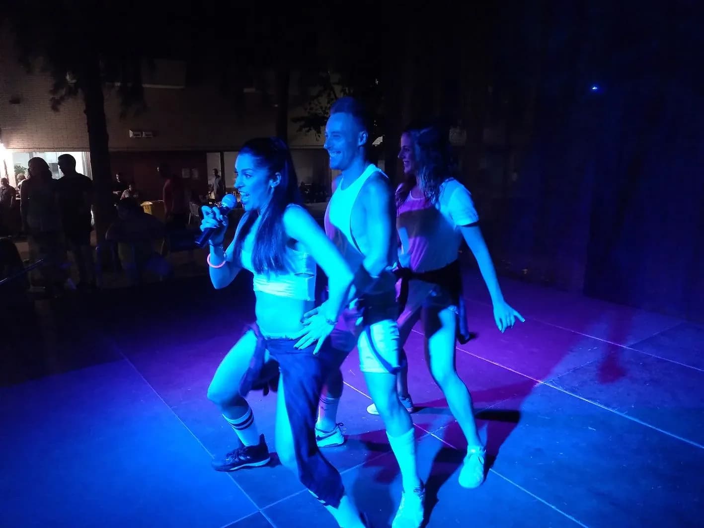 Animadora cantando con microfono junto a bailarines en pista al aire libre con iluminacion azul y morada