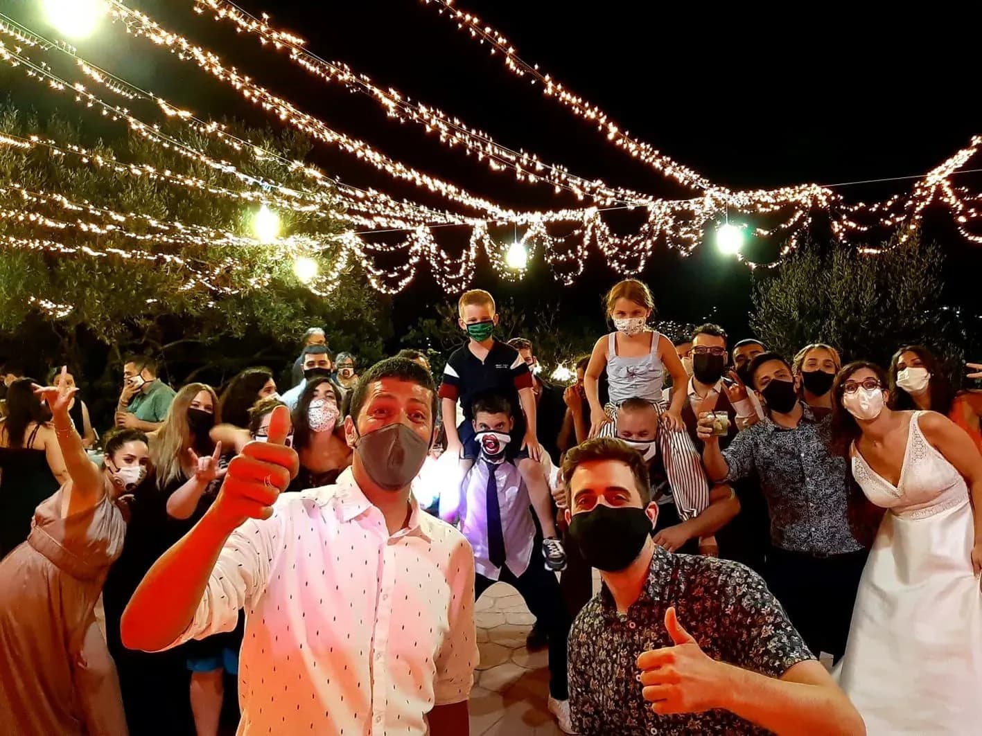 Novios con mascarilla y invitados haciéndose selfie de grupo bajo guirnaldas de bombillas en boda al aire libre de noche