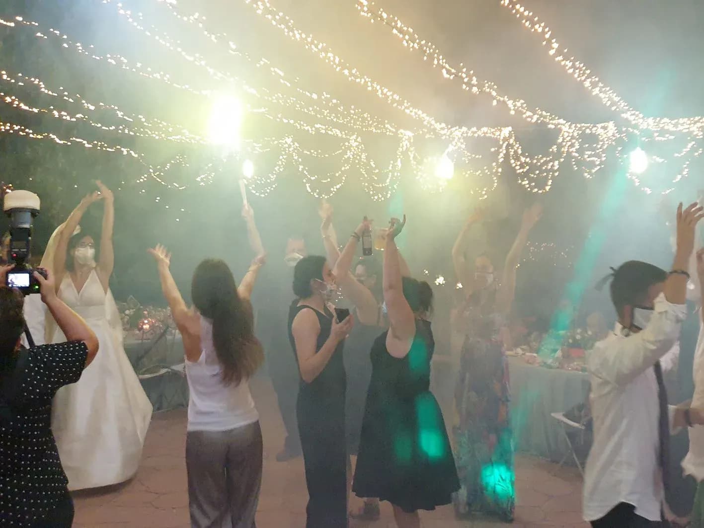 Novia y invitados con mascarilla bailando con los brazos en alto bajo cortinas de luces de hadas en boda nocturna