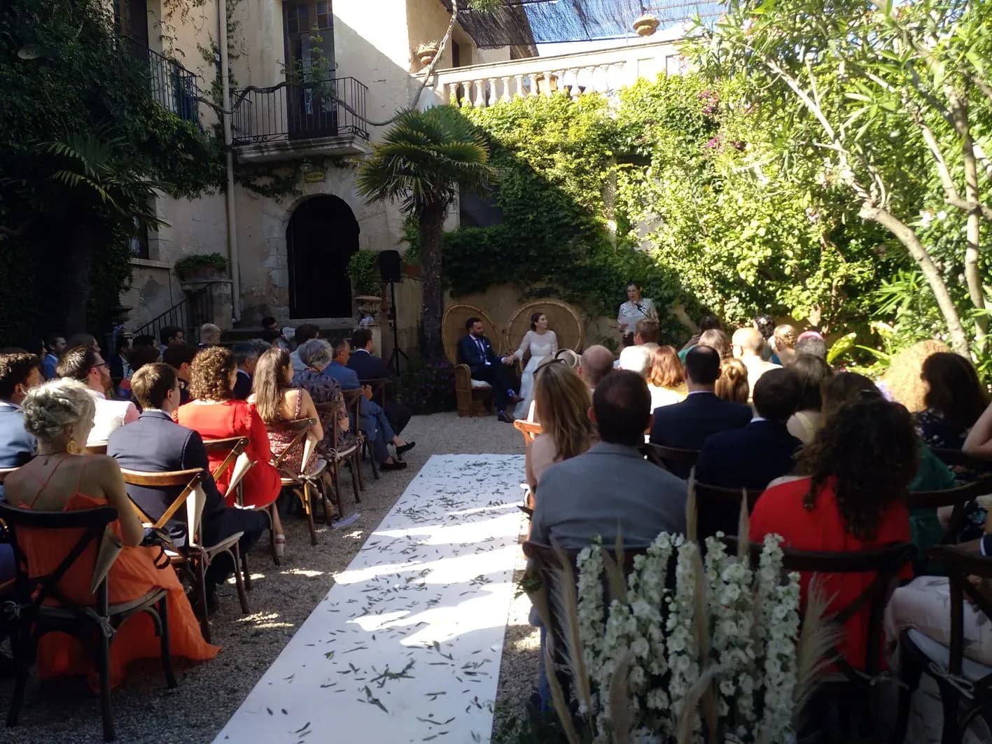 Ceremonia civil de boda en patio interior con hierba al fondo, novios sentados frente a invitados y camino de pétalos blancos