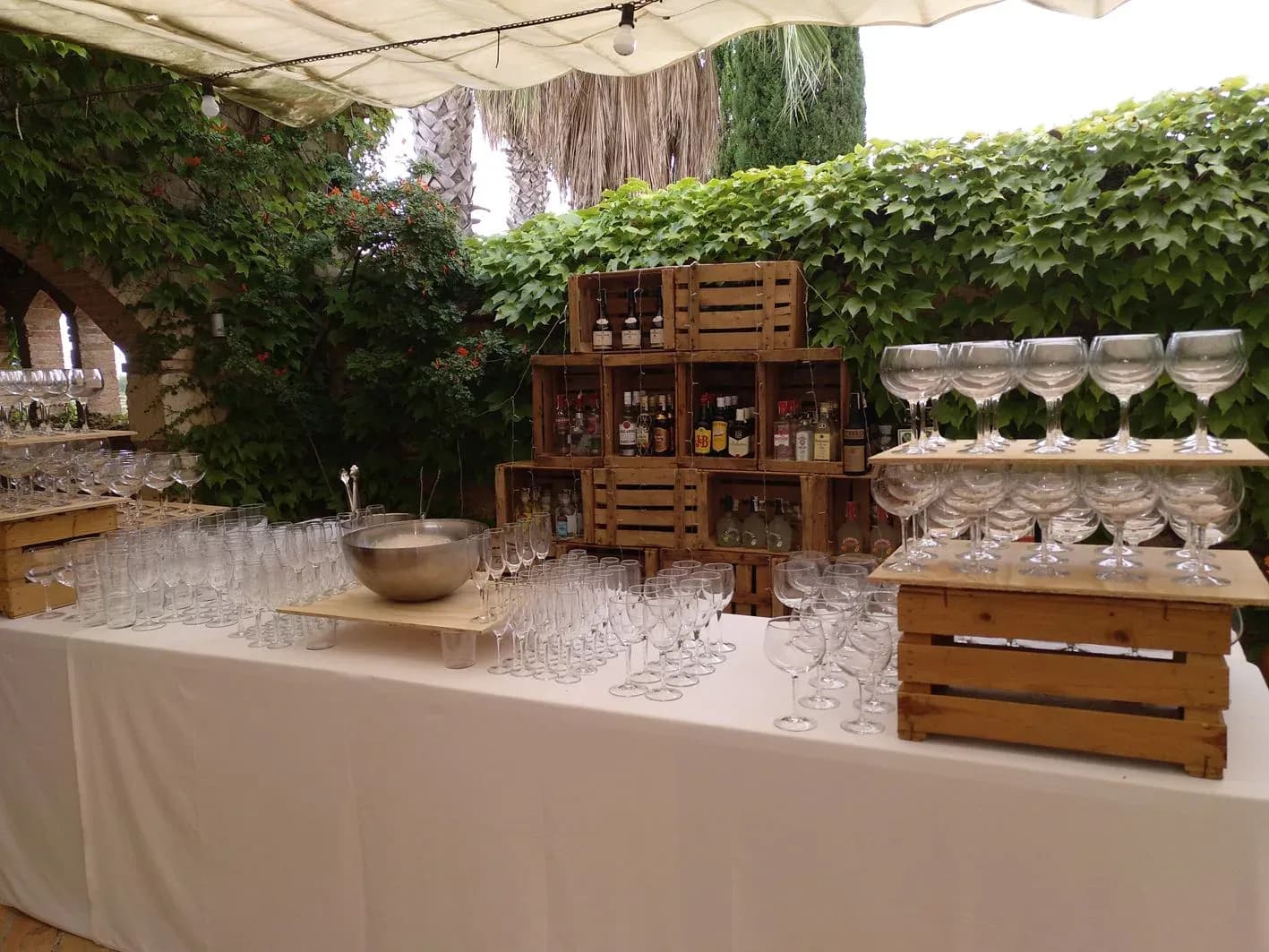 Barra de coctel para boda con copas de balon alineadas, botellas en cajas de madera y muro vegetal de hiedra detras
