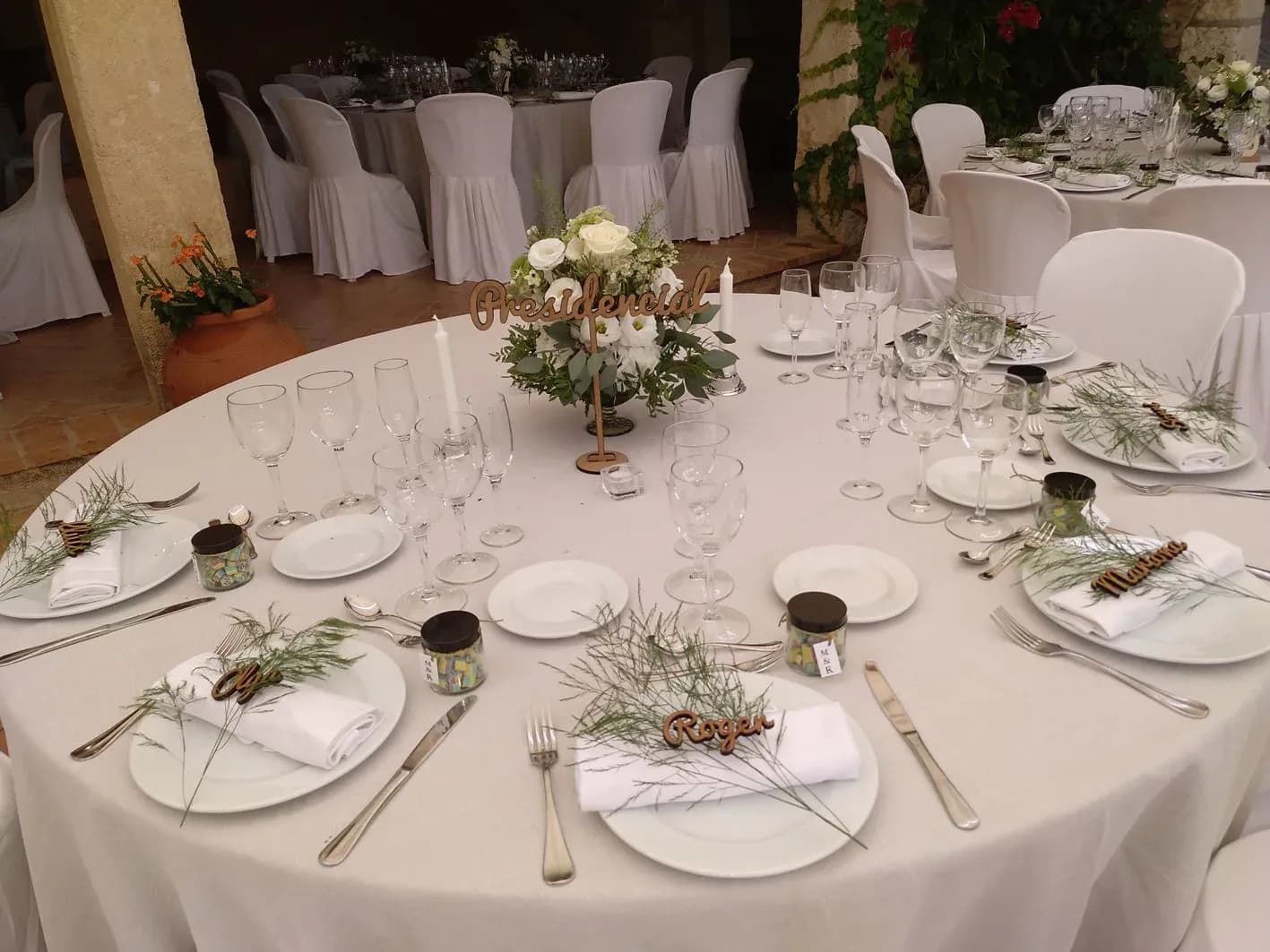 Mesa presidencial de boda con sillas blancas, centro floral, nombres tallados en madera y cubertura de plata