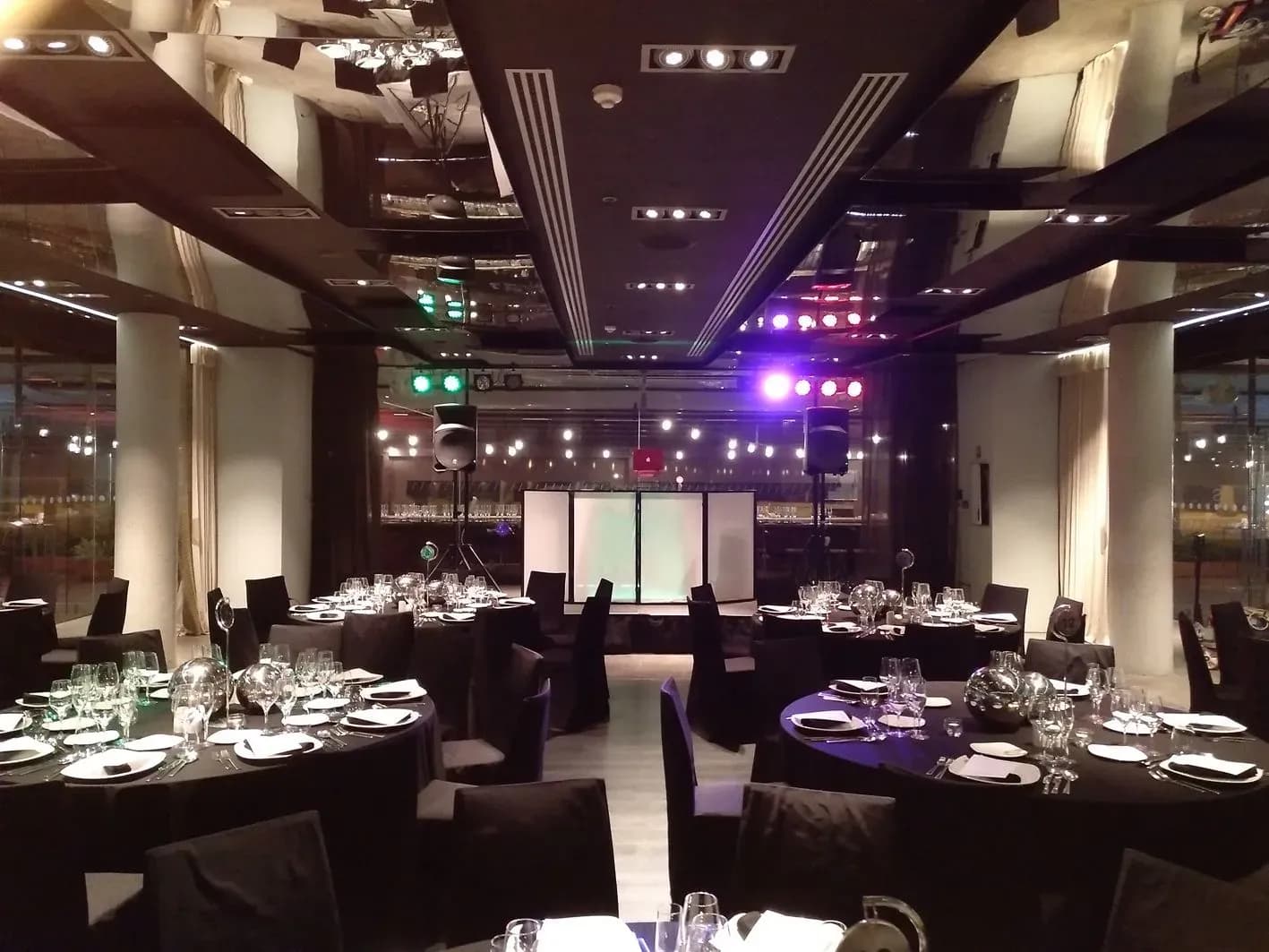 Salon de bodas montado con mesas redondas vestidas, altavoces y pantalla central iluminada en violeta