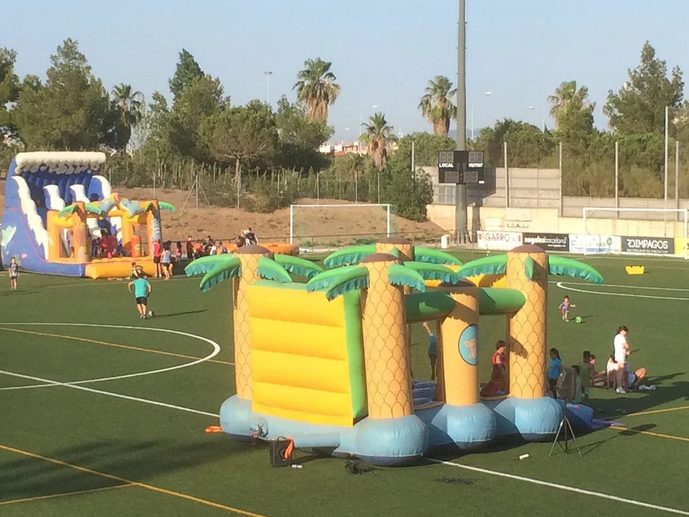 Castillo hinchable amarillo con palmeras y tobogán en campo de fútbol con torre de luz y otro hinchable azul al fondo