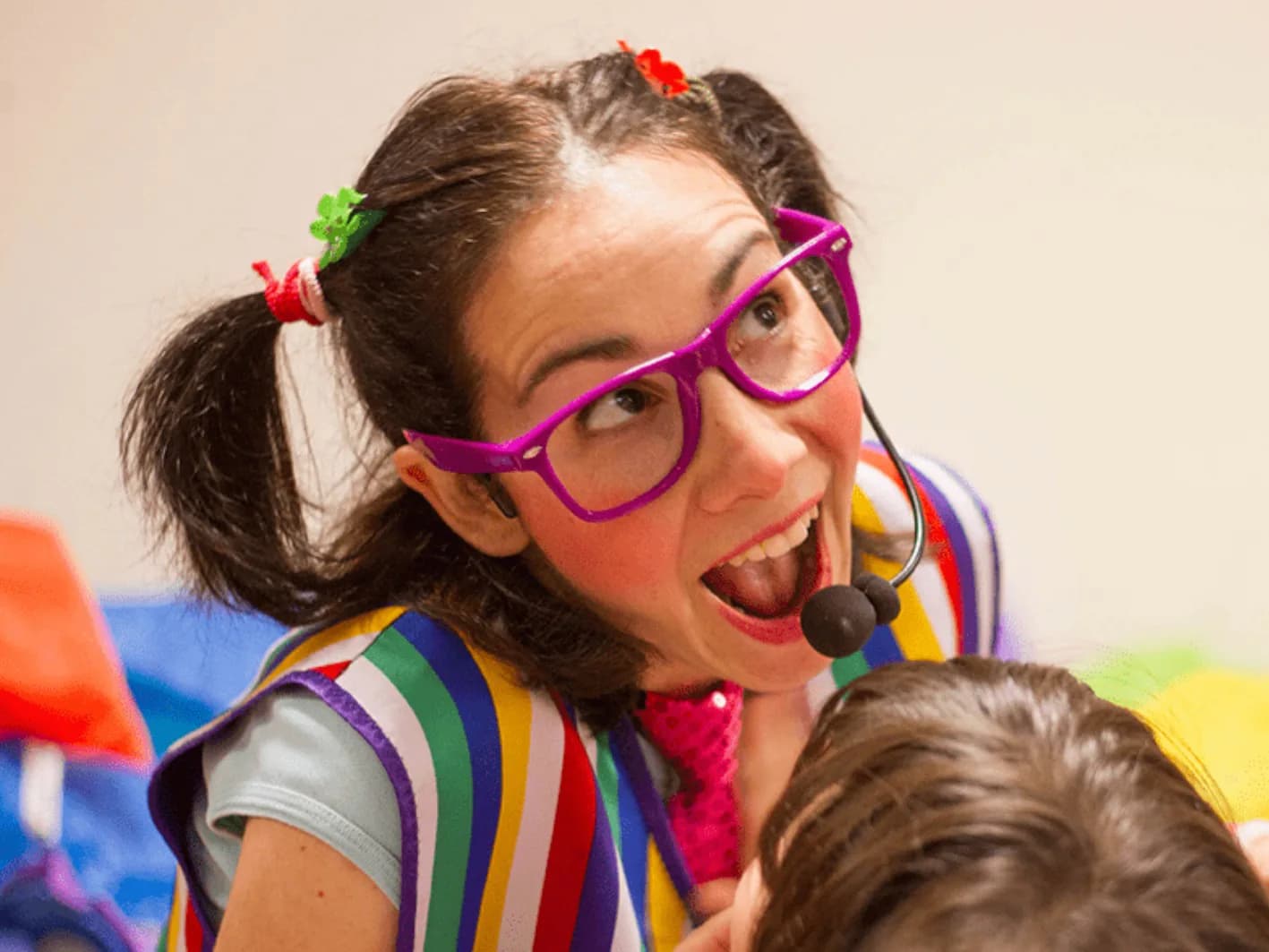 Jessica con gafas violetas, coletas y vestido a rayas multicolor sonriendo a un niño durante animación infantil