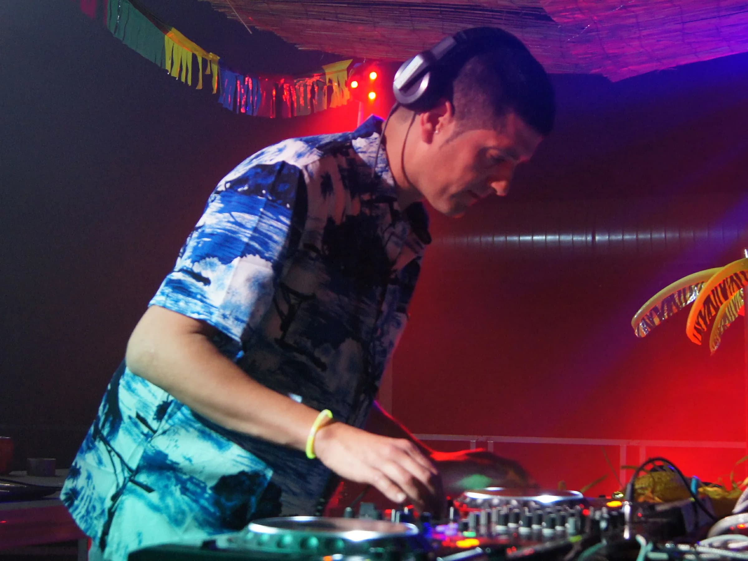 DJ con camisa hawaiana y auriculares pinchando bajo guirnaldas de colores y techo de cañizo en fiesta tropical de verano