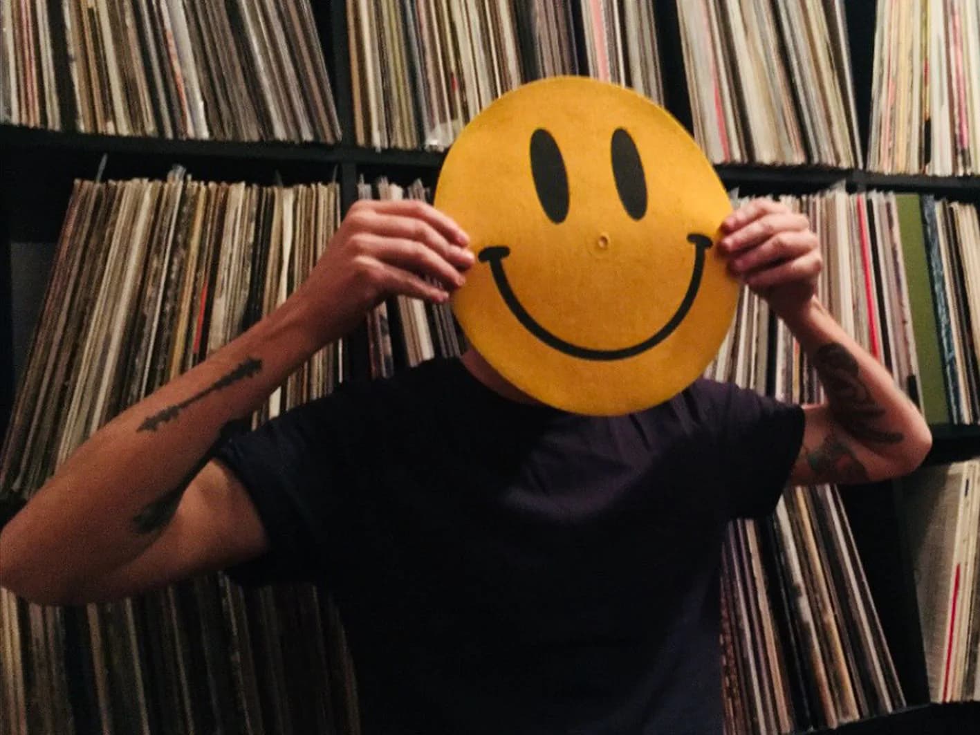 Persona cubriéndose la cara con slipmat smiley amarillo frente a estantería repleta de vinilos, imagen del catálogo electrónica